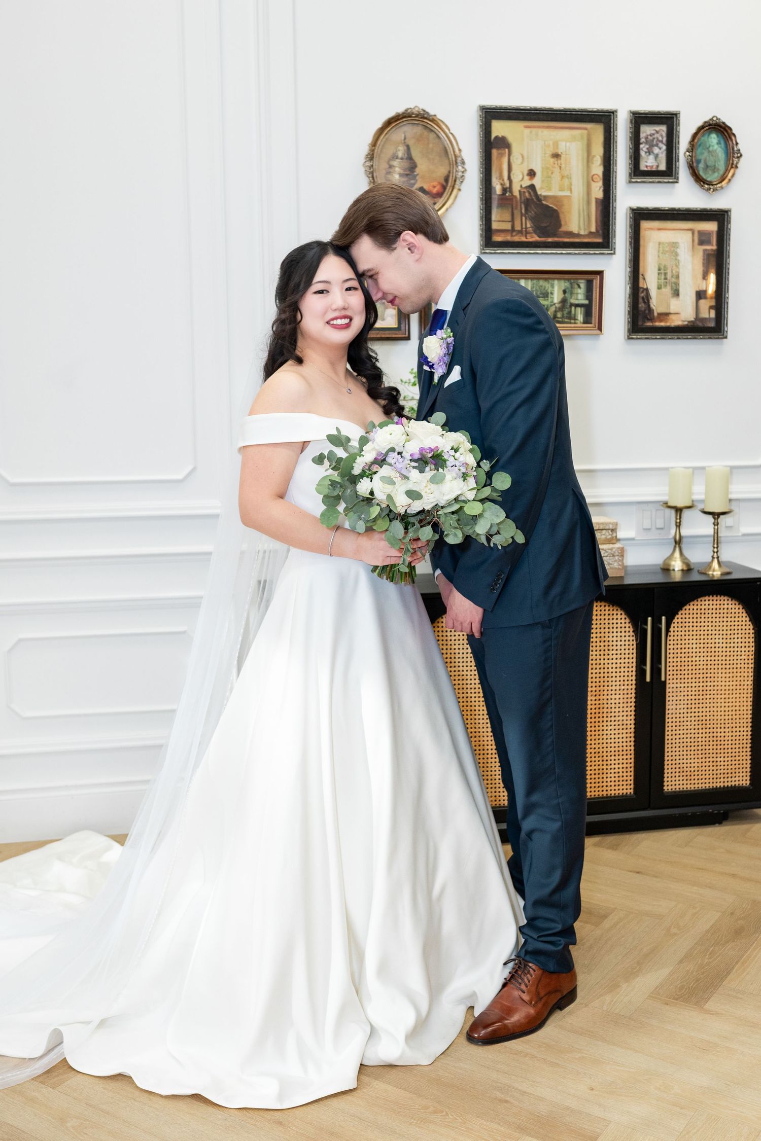 A winter wedding scene with a bride in a flowing white gown and groom in dark suit standing in snow-covered woods.