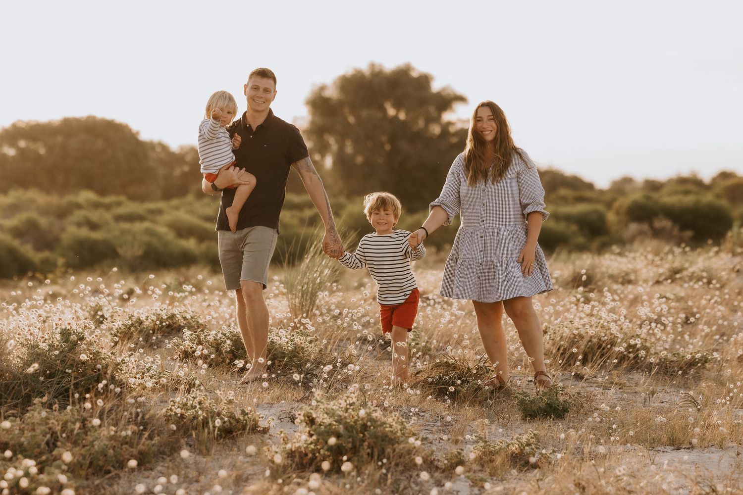 Family of four walking through a sunny wildflower field, parents holding toddler hands at golden hour.