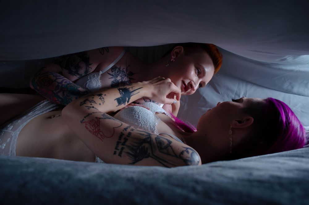 Two people with tattoos sharing an intimate moment under dim lighting on a bed.