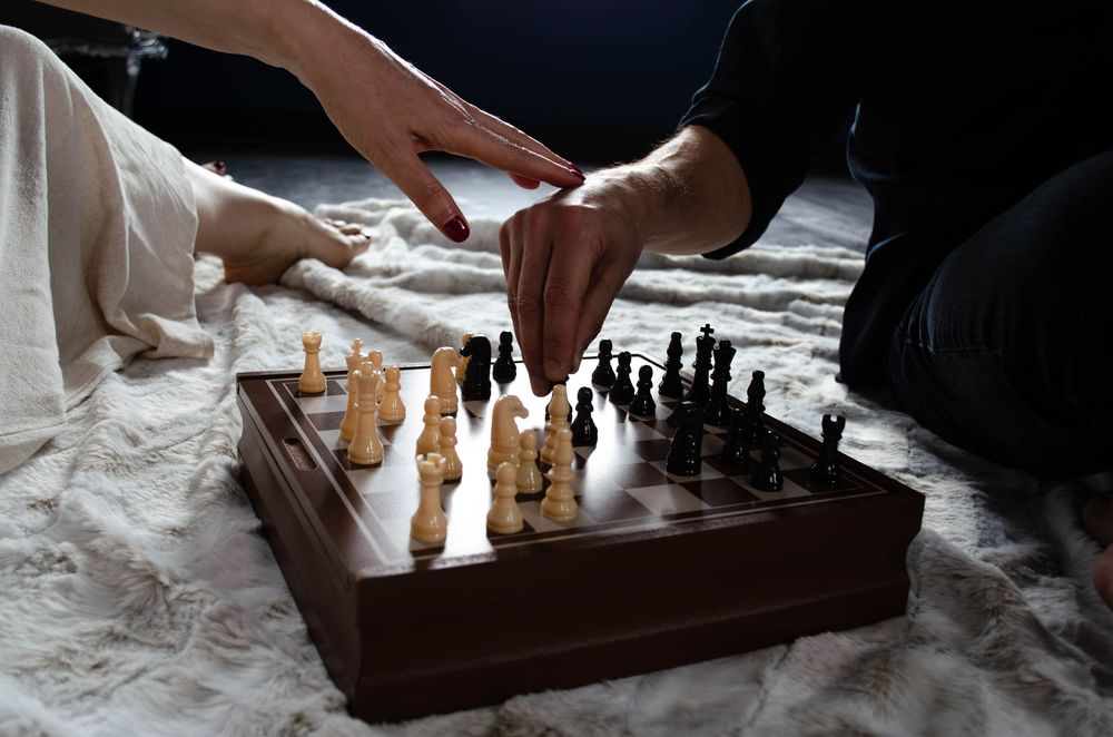 Two people play a game of chess with wooden pieces on a wooden board in dramatic lighting.