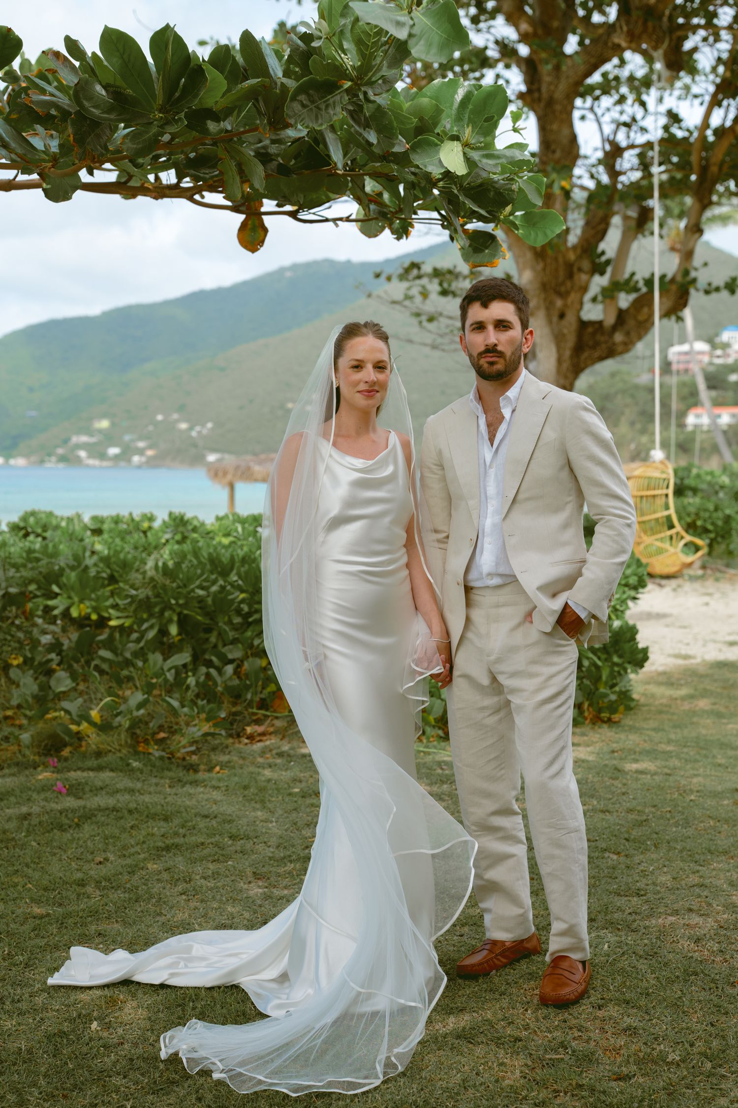 A couple poses for wedding photos on a scenic tropical beach with mountains in the background.