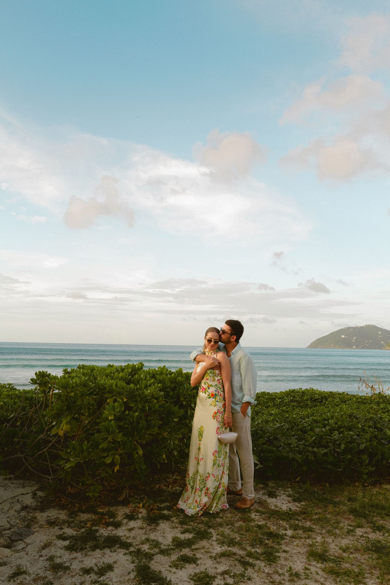 A couple poses for wedding photos on a scenic tropical beach with mountains in the background.