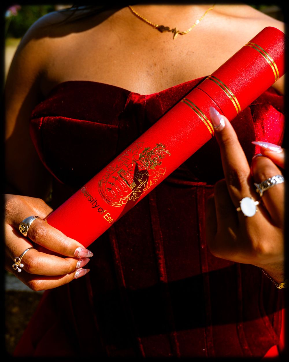 Hands wearing rings hold a bright red decorative scroll or diploma against a burgundy velvet dress background.