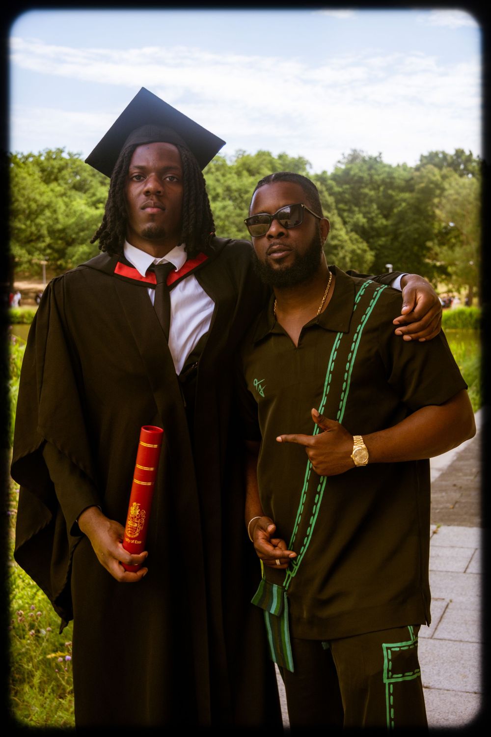 Two friends in graduation attire pose together while holding colorful light-up bottles in an outdoor setting.