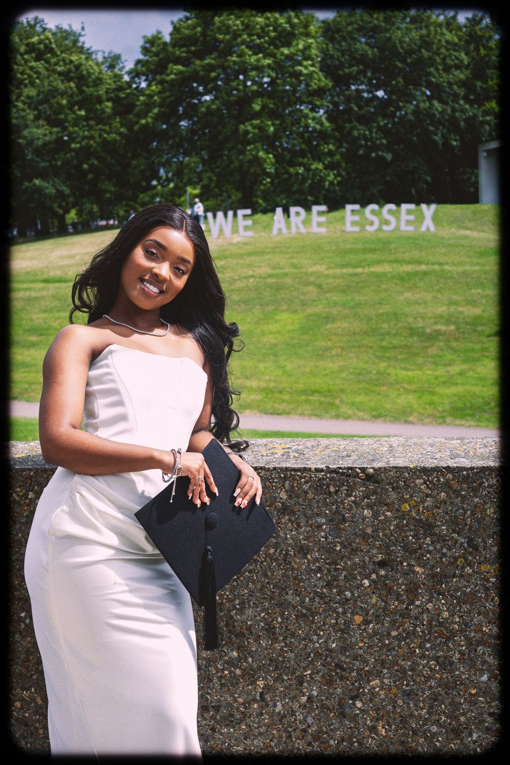 Someone in a white dress poses in front of a 'WE ARE ESSEX' sign on a grassy hill on a sunny day.