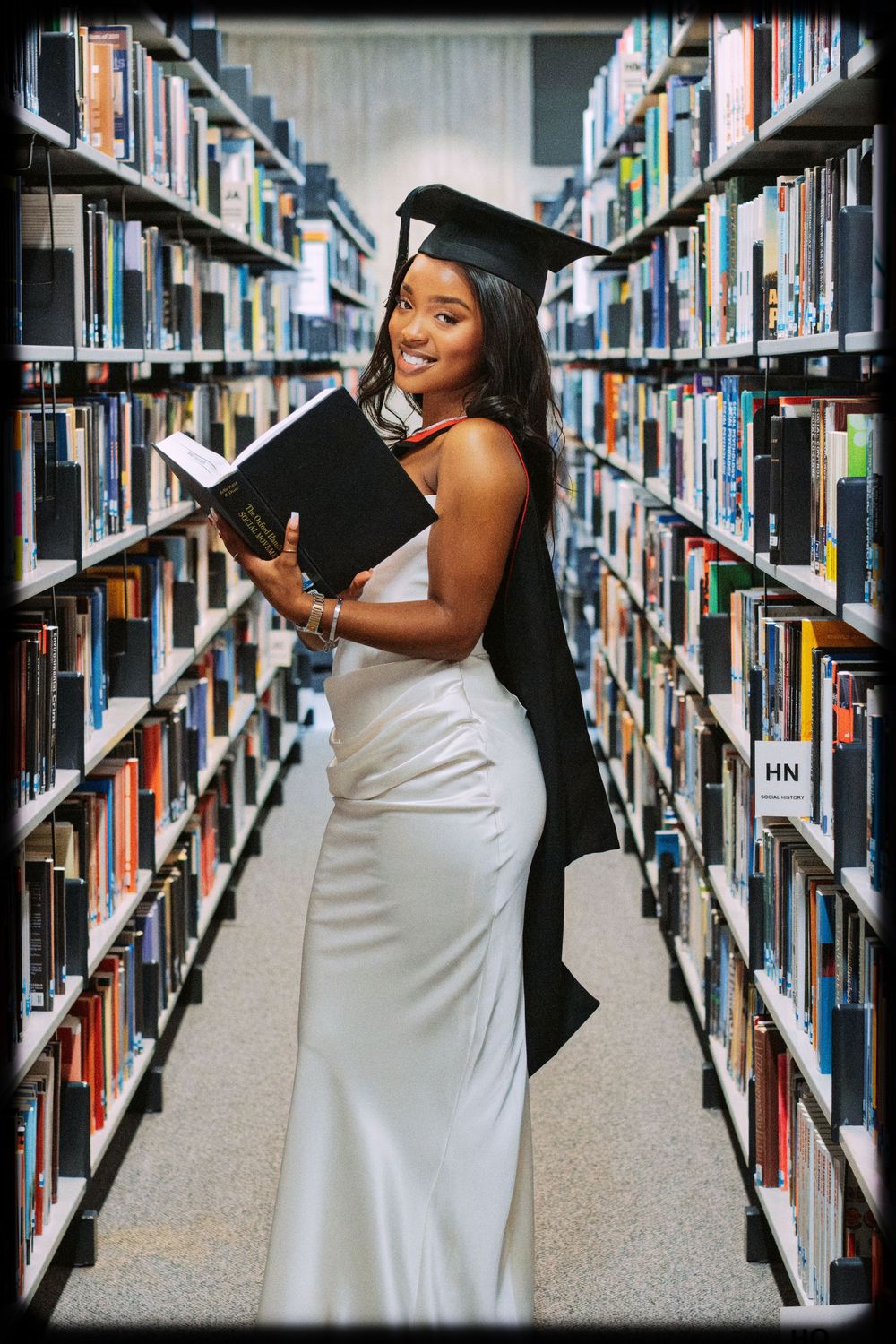 Student in white dress browses books between library shelves while posing playfully in a sequence of photos.