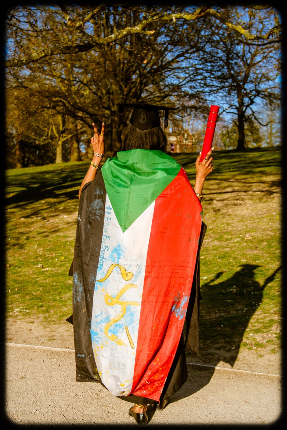 A person draped in a Sudanese flag stands on a path in a sunny park with autumn trees in the background.