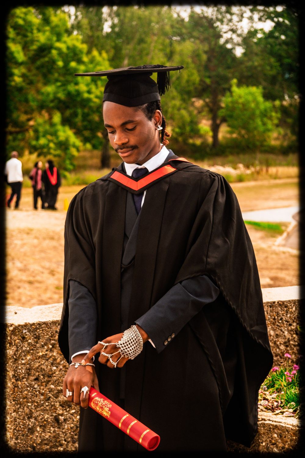 Graduate in cap and gown smiling while holding diploma during outdoor commencement ceremony.