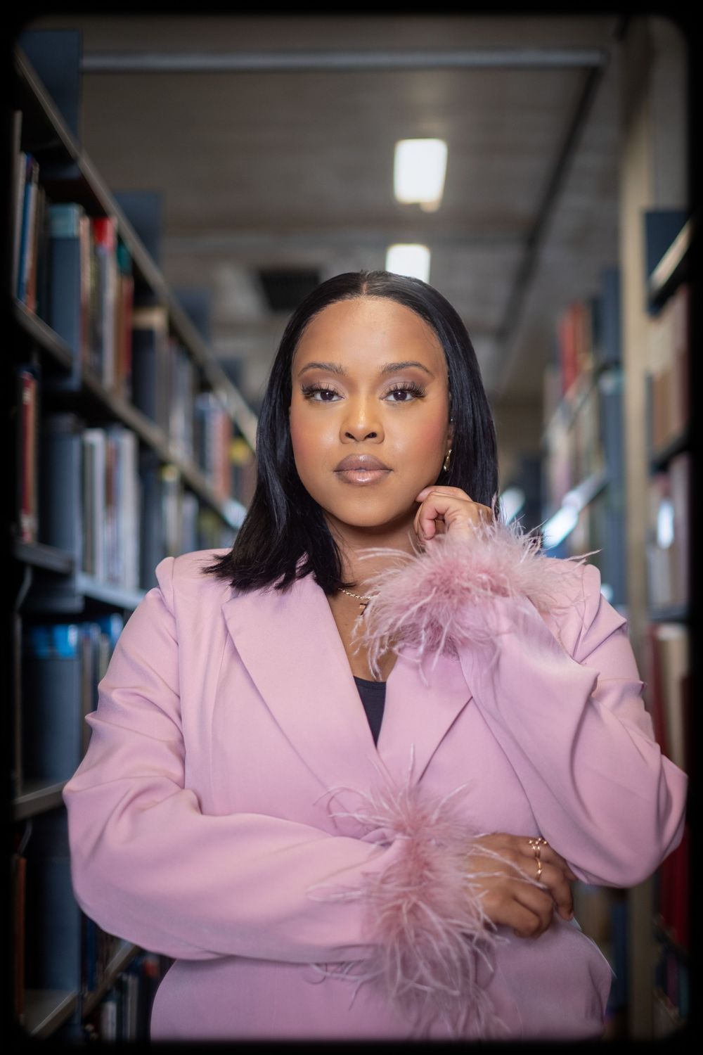 Professional portrait series in pink blazer with feather detail against library bookshelves background.