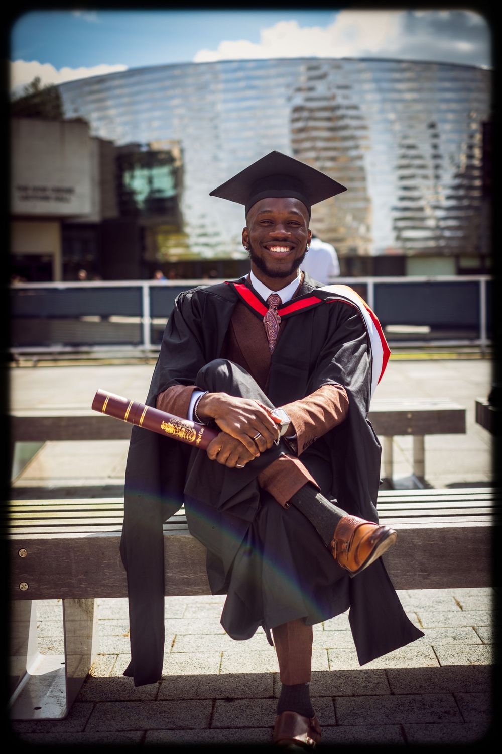 A graduate in cap and gown poses with their diploma on outdoor steps in an urban setting.