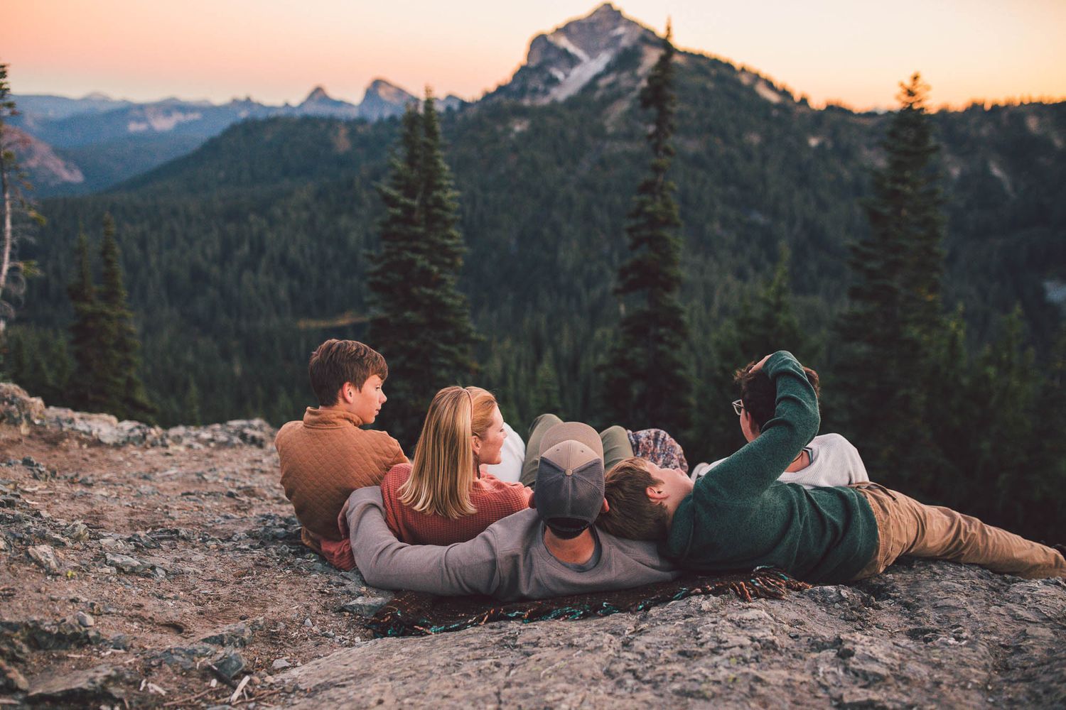 Family lay on a rocky mountain ledge watching a colorful sunset over evergreen forest peaks in the distance in Mt. Rainier National Park