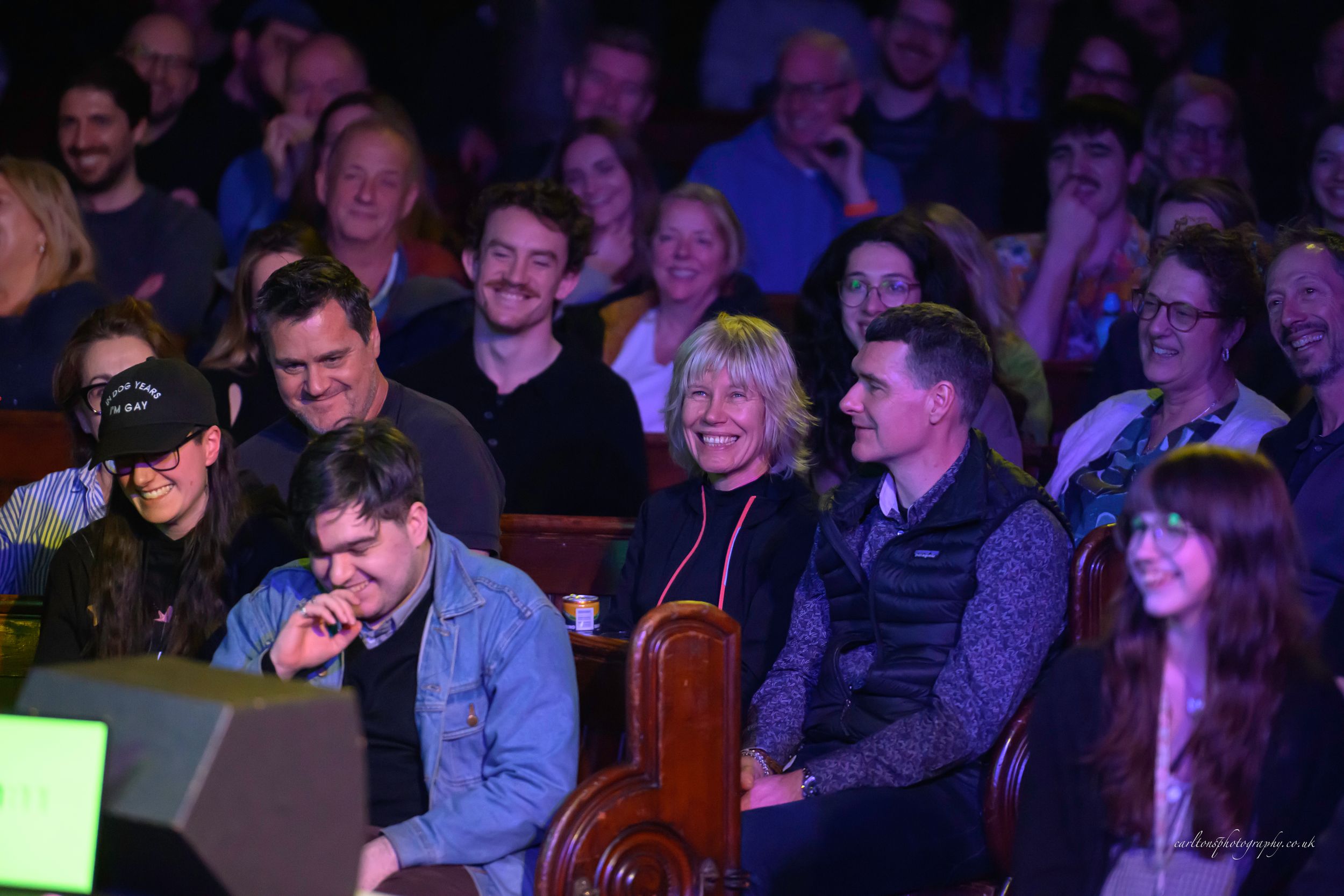 An audience in a dark theater watches intently with smiles and laughter illuminated by purple stage lighting.