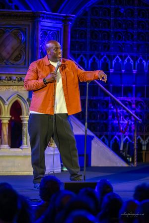 Performer in an orange jacket gestures energetically on stage with dramatic blue lighting and gothic architecture behind.
