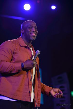 Stand-up comedian in orange jacket performs against dark background with blue stage lighting.