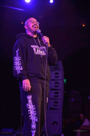 A performer in a black outfit with glowing purple cross designs stands on stage under blue lighting.