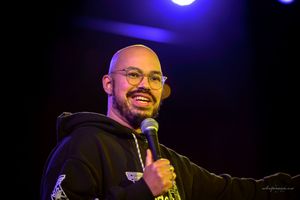 A performer on stage smiling and holding a microphone under blue stage lighting during a comedy show.