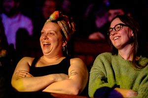 Theater audience members react with joy and amusement while bathed in purple and blue stage lighting.