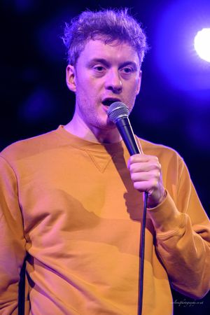 A performer in a yellow sweater sings into a microphone on stage with purple lighting in the background.