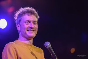 A performer in casual attire gestures expressively while speaking into a microphone under theatrical lighting.