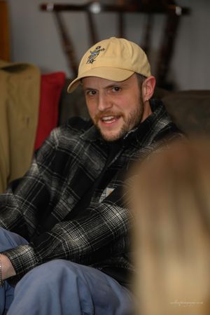 A person wearing a plaid flannel shirt and baseball cap sits casually indoors.