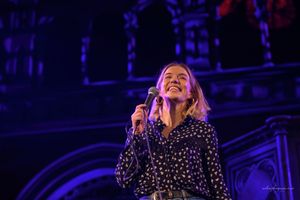 A performer in a polka dot dress sings passionately on stage under dramatic blue lighting during a concert performance.