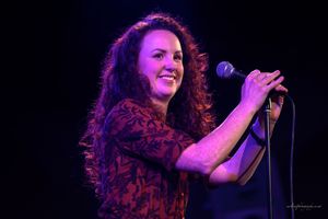 A performer with curly hair sings passionately into a microphone on a purple-lit stage.