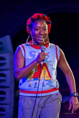 A performer in a striped outfit and white tank top smiles while holding a microphone on stage with colorful lighting.