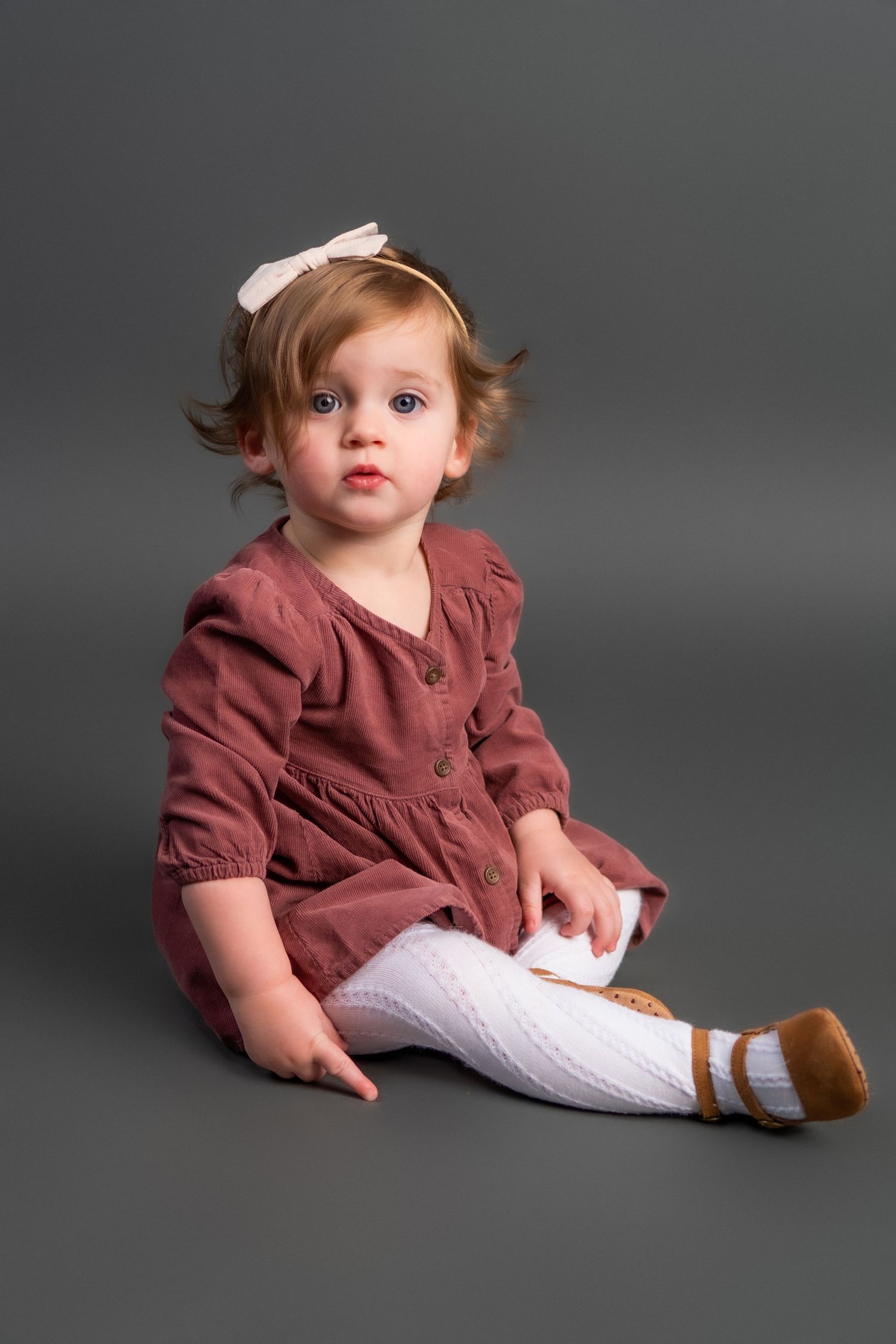 Baby wearing burgundy dress and white tights sits on gray backdrop in professional studio portraits.