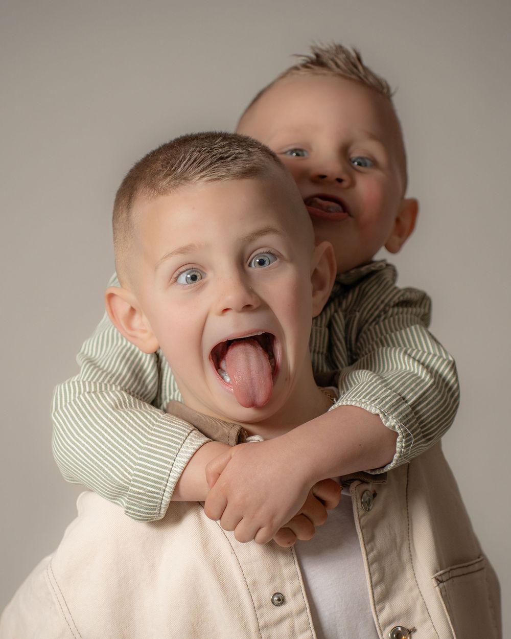 Two young children in striped and light colored clothing making silly faces and hugging while posing for a portrait.