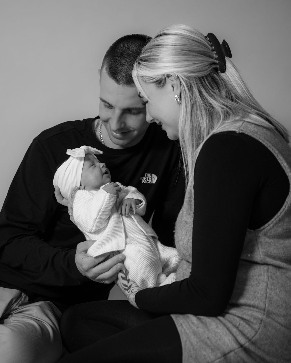 A black and white portrait of parents sharing an intimate moment with their newborn baby wearing a white bow headband.