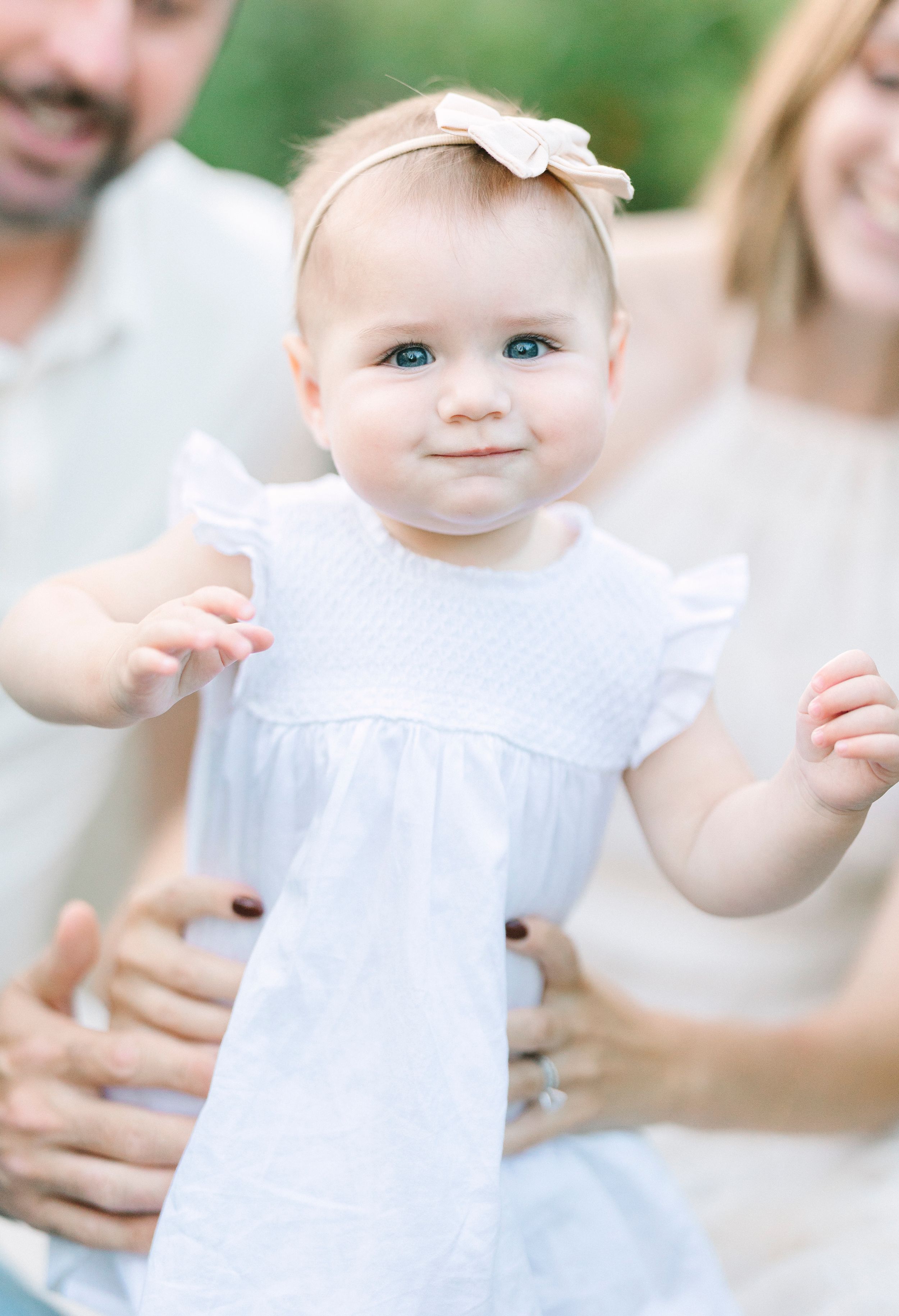 Baby in white dress against soft outdoor background with gentle lighting.