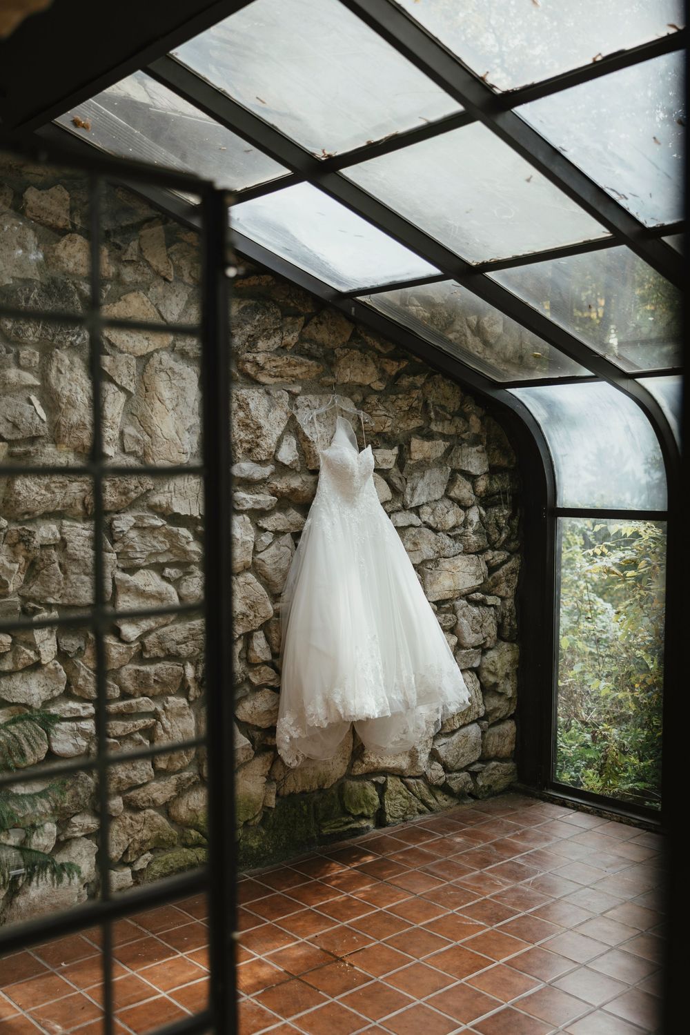 A white wedding dress hangs elegantly in a sunlit stone conservatory with terracotta flooring and rustic walls.