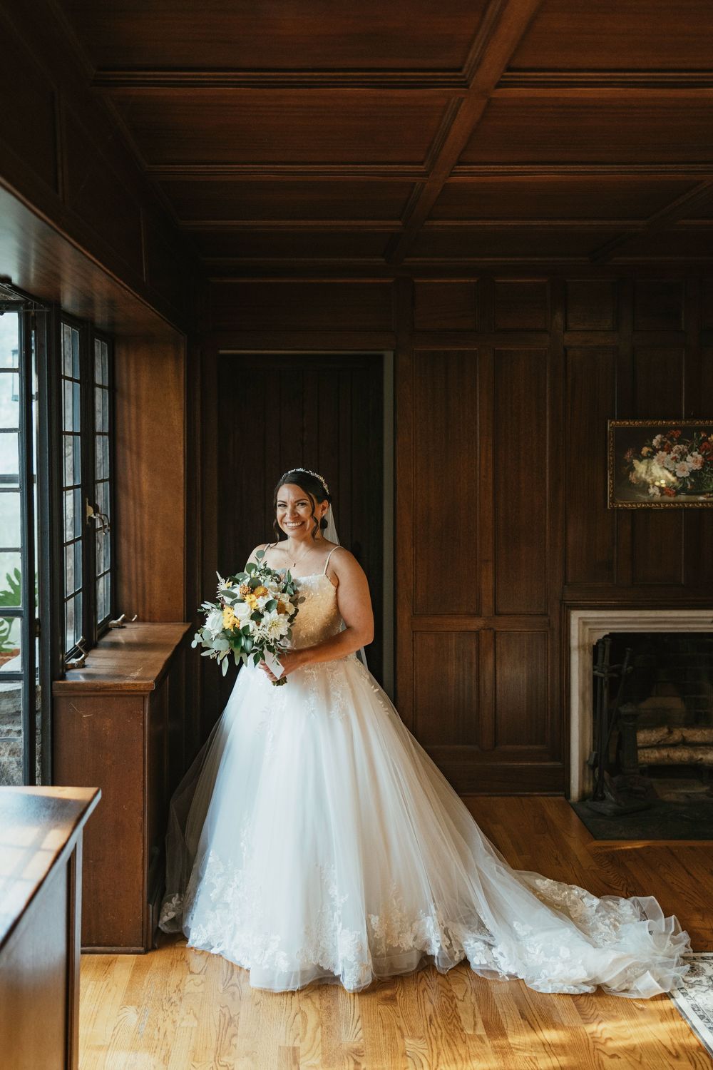 An elegant ballgown wedding dress with tulle skirt catches sunlight through wooden-paneled room windows.
