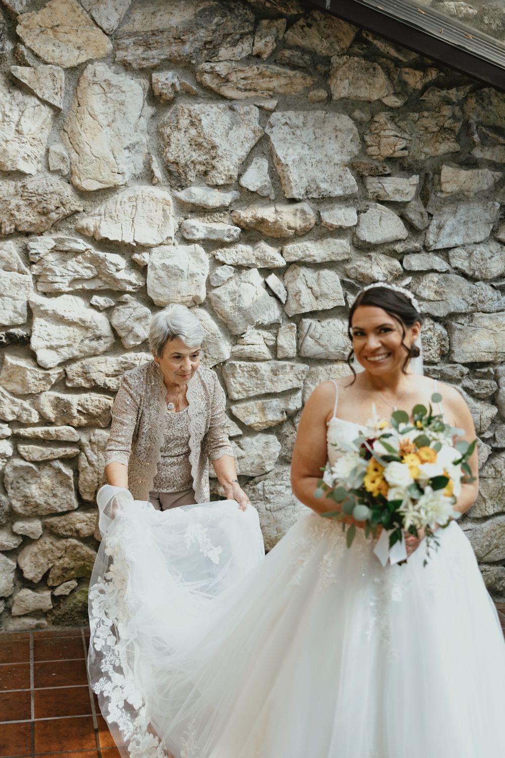 A bride in a white lace wedding gown sits on rustic stone steps holding a vibrant sunflower and white floral bouquet.