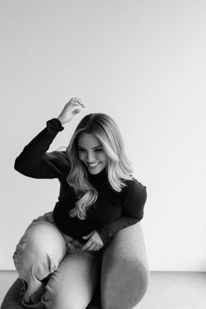 Black and white portrait of someone in a dark sweater sitting and laughing joyfully against a white wall.