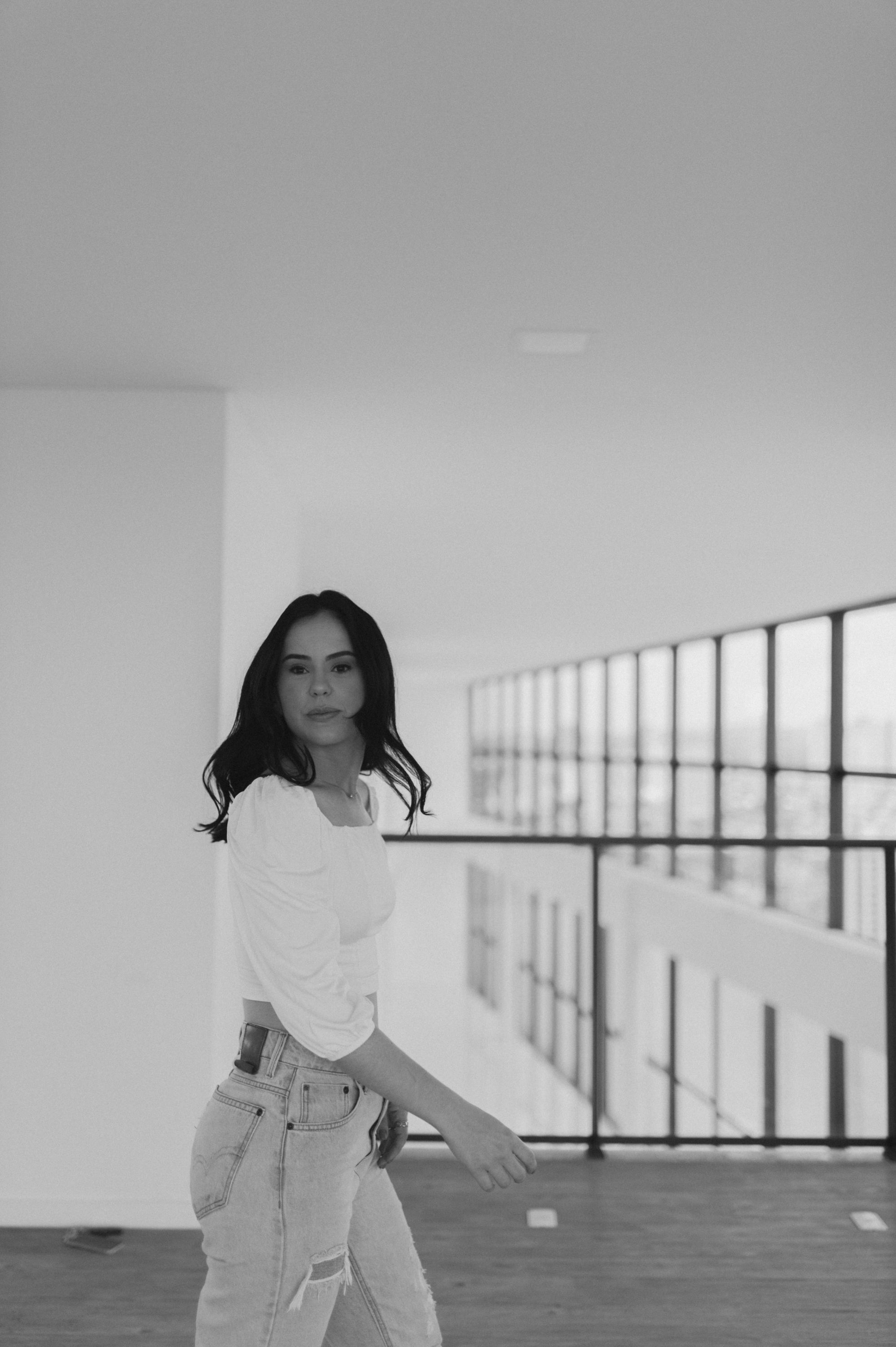 Black and white photo of a person in white top and distressed jeans standing in a minimalist space.