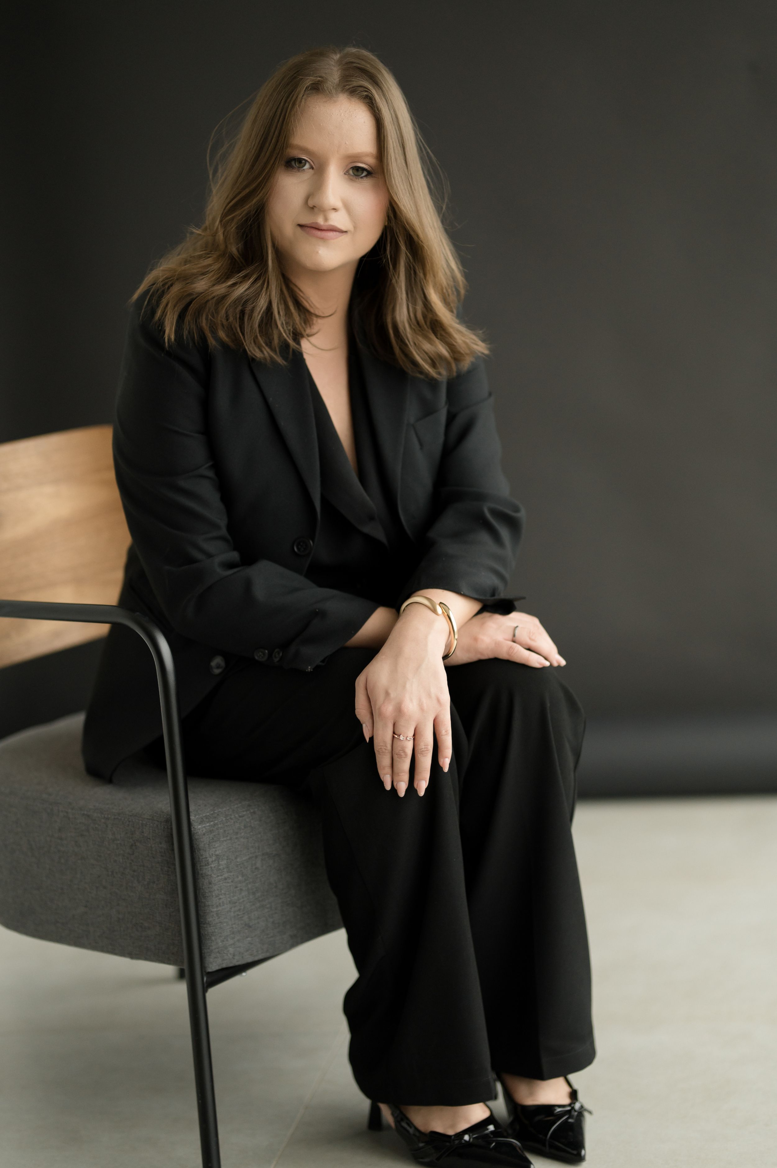 Professional studio portrait of someone in a black business suit seated in a modern chair against a dark background.