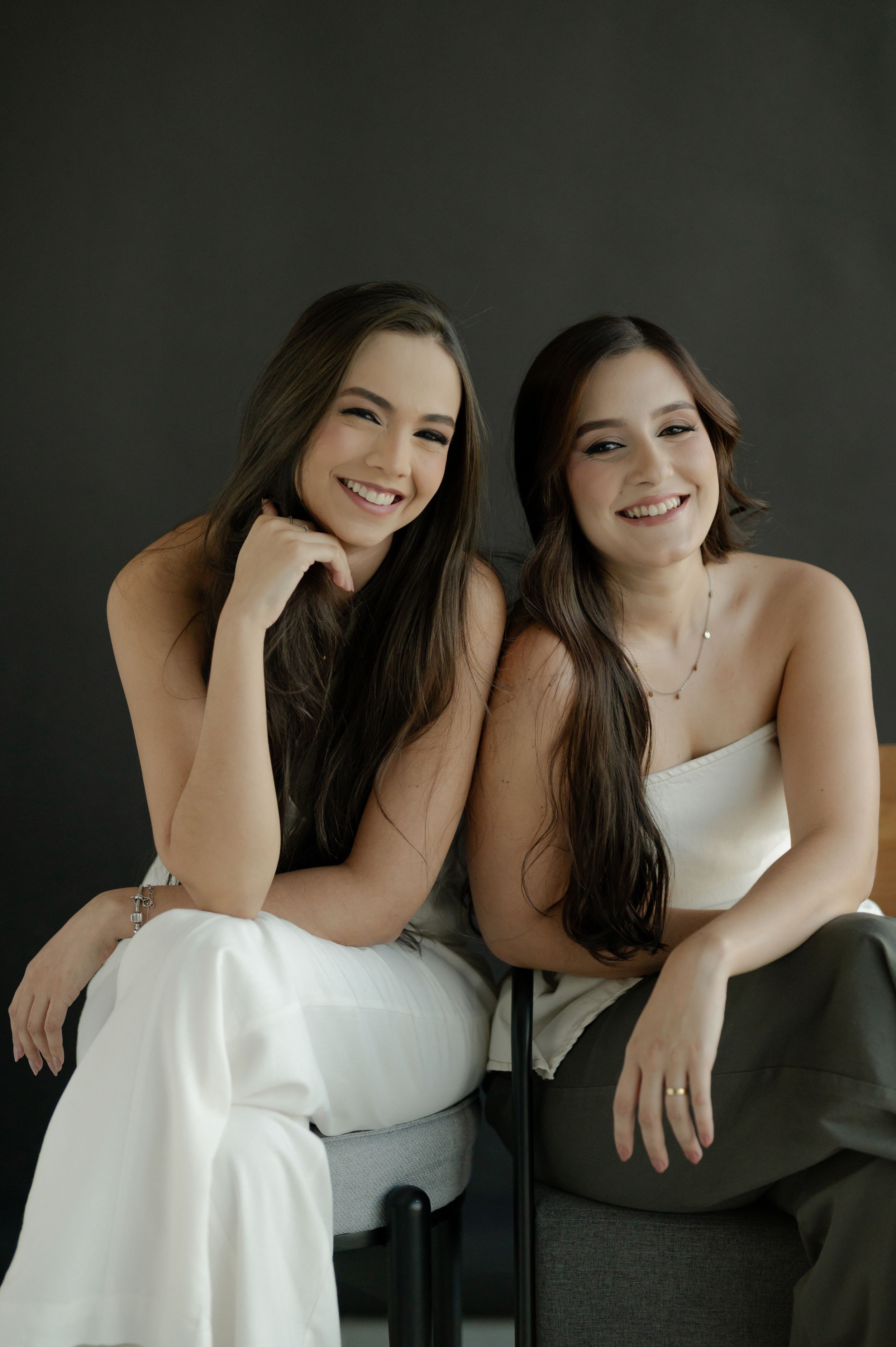 Two friends in contrasting black and white outfits share candid laughs during a studio photoshoot against dark backdrop.