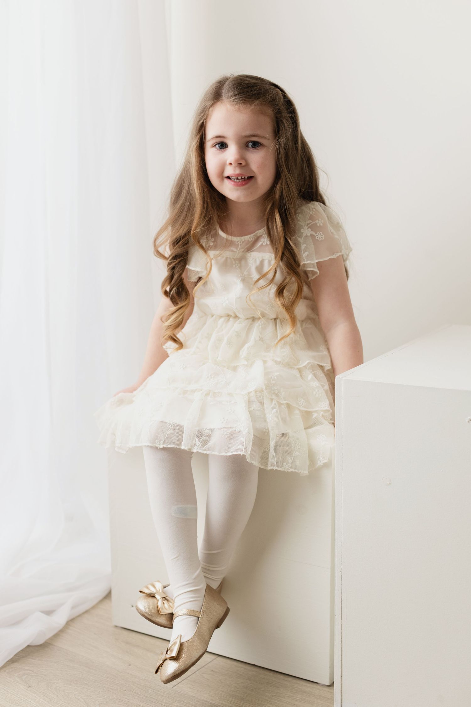 A young child in a cream-colored ruffled dress and white leggings poses against a light background.