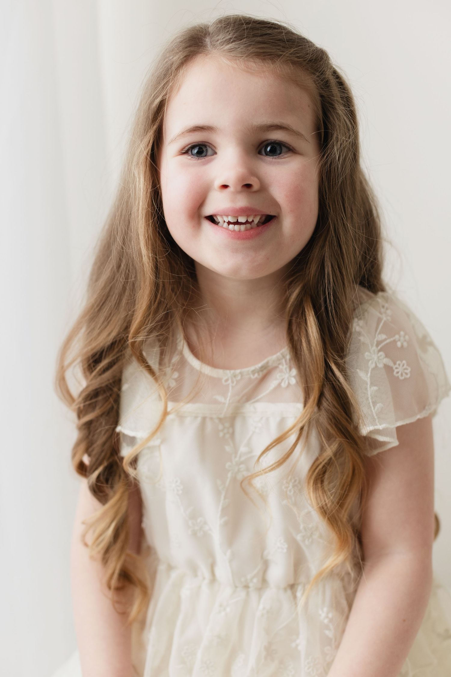 Young child wearing a cream-colored lace dress with ruffled sleeves and long wavy brown hair smiles against white background.