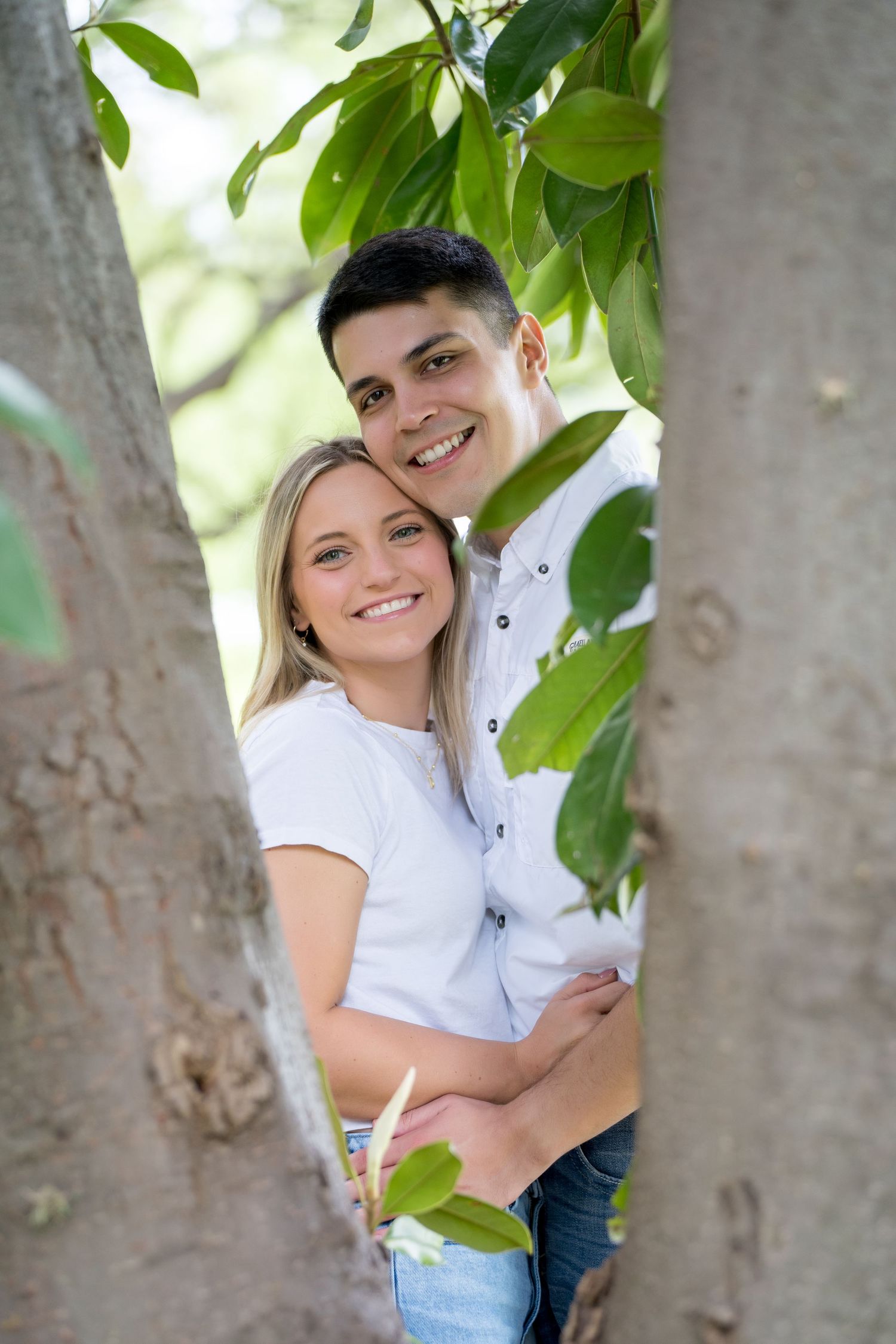 Happy young couple embracing among green tree branches in a sunny park, both wearing white shirts.