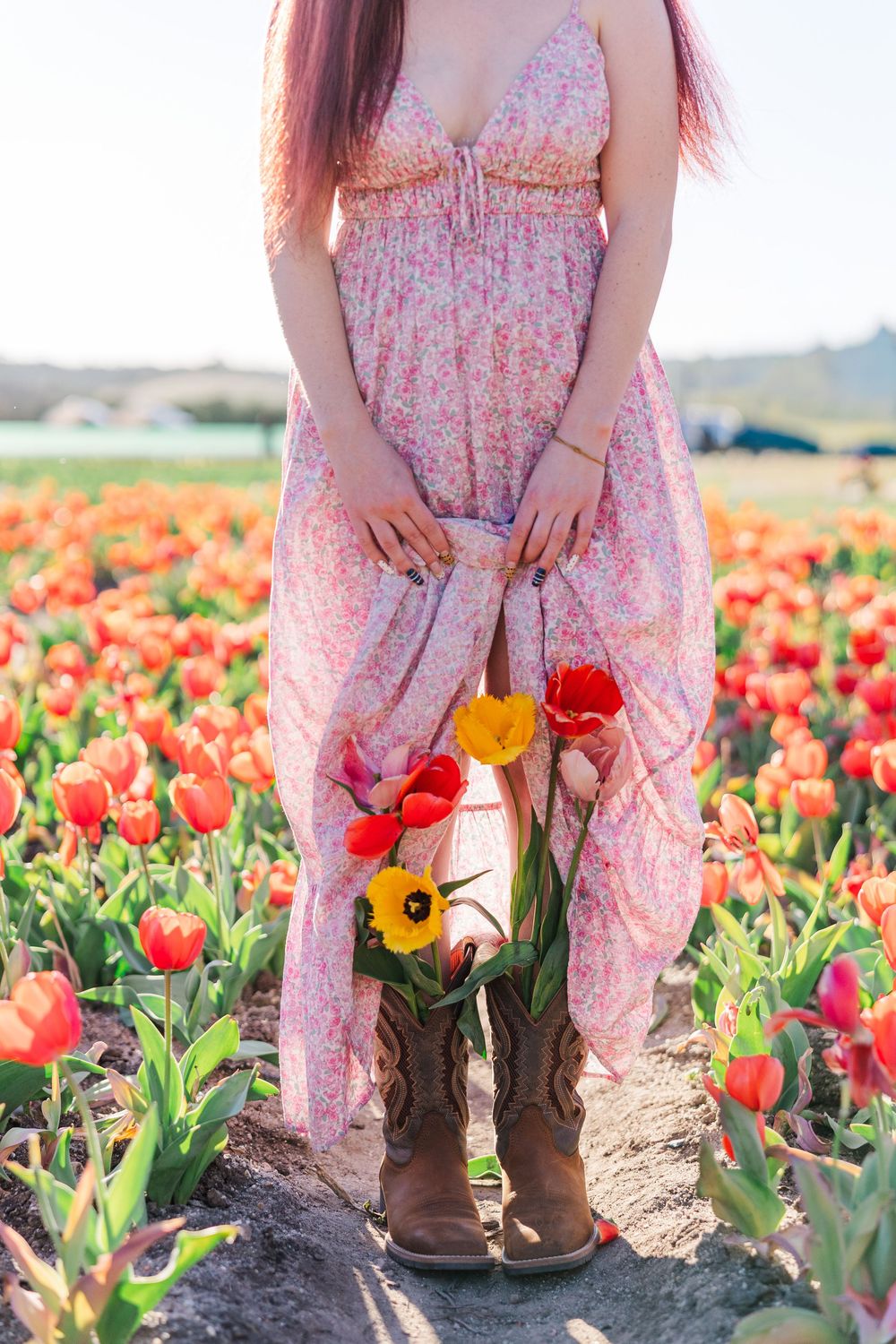 Brown boots stand among vibrant red and yellow tulips in a sunny flower field against a soft pink floral dress.