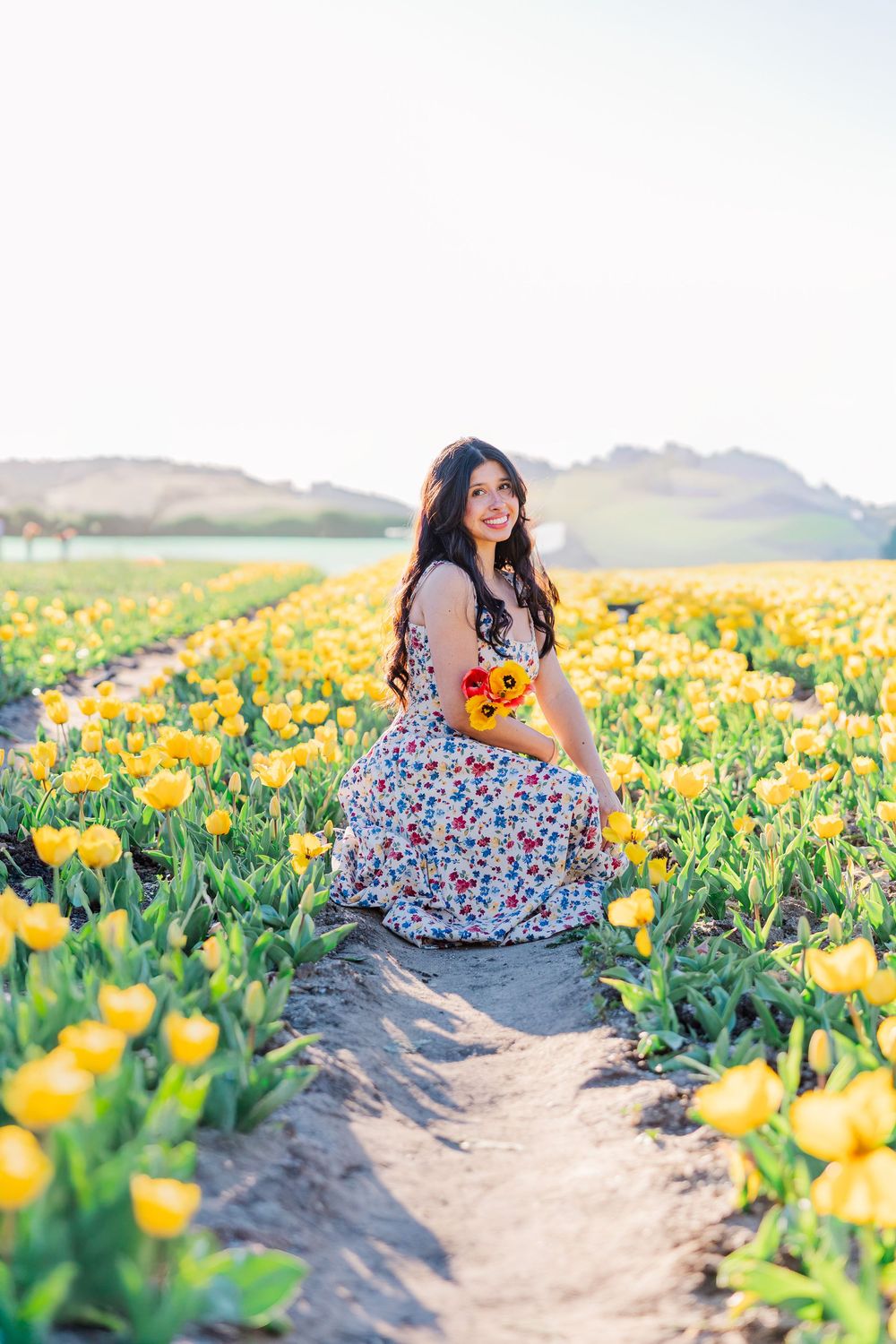A floral dress-clad figure sits among vibrant yellow tulip fields on a sunny spring day.