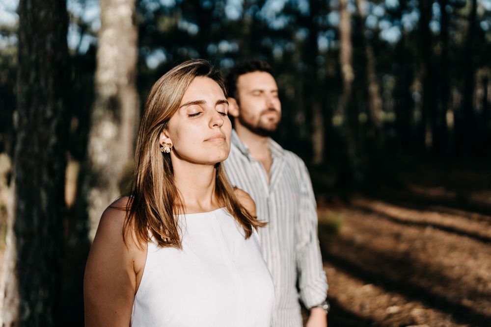 A couple stands in a sunlit forest, with warm light filtering through the trees as they pose with closed eyes.