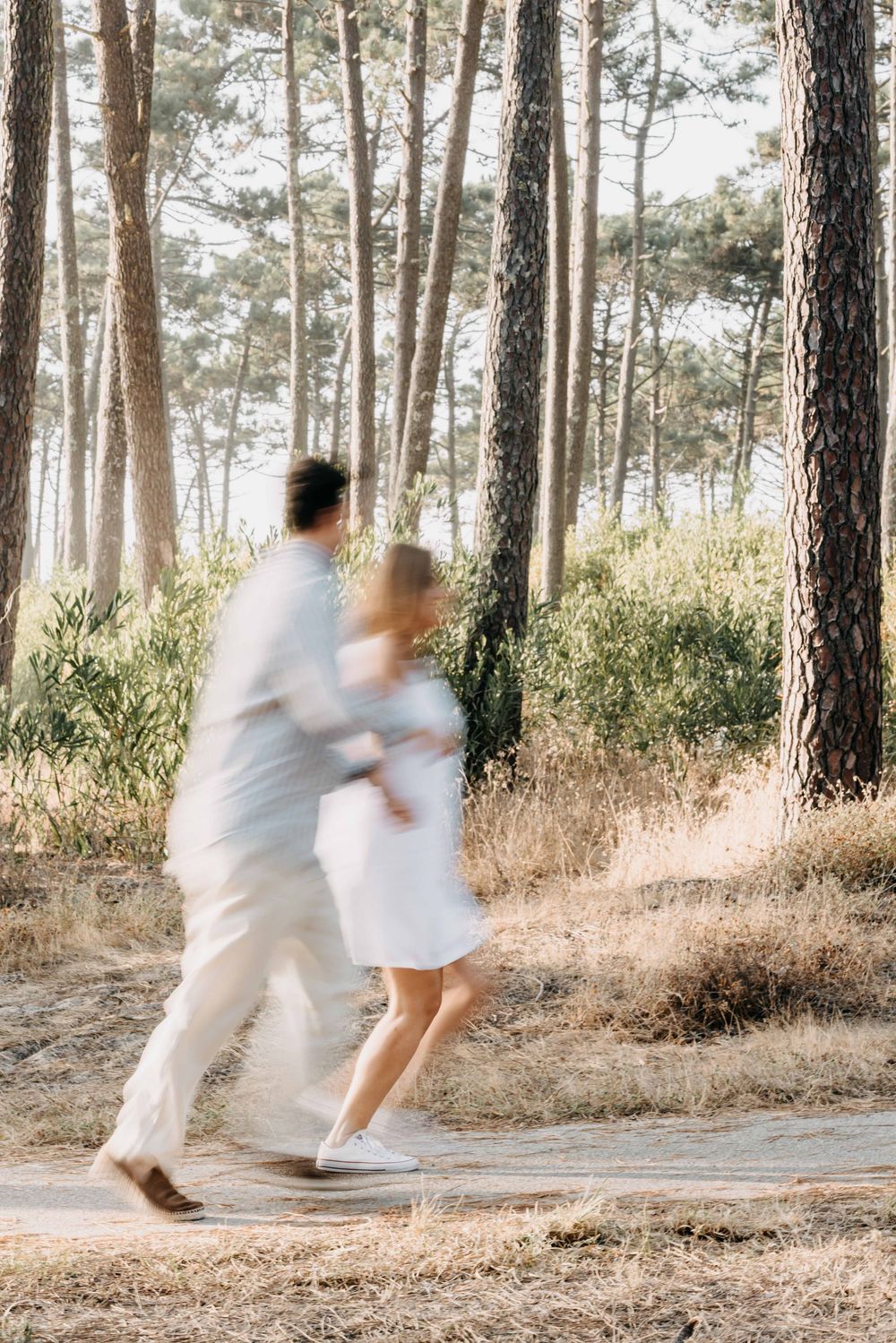 A couple in white outfits playfully spins and dances together in a sunlit pine forest.