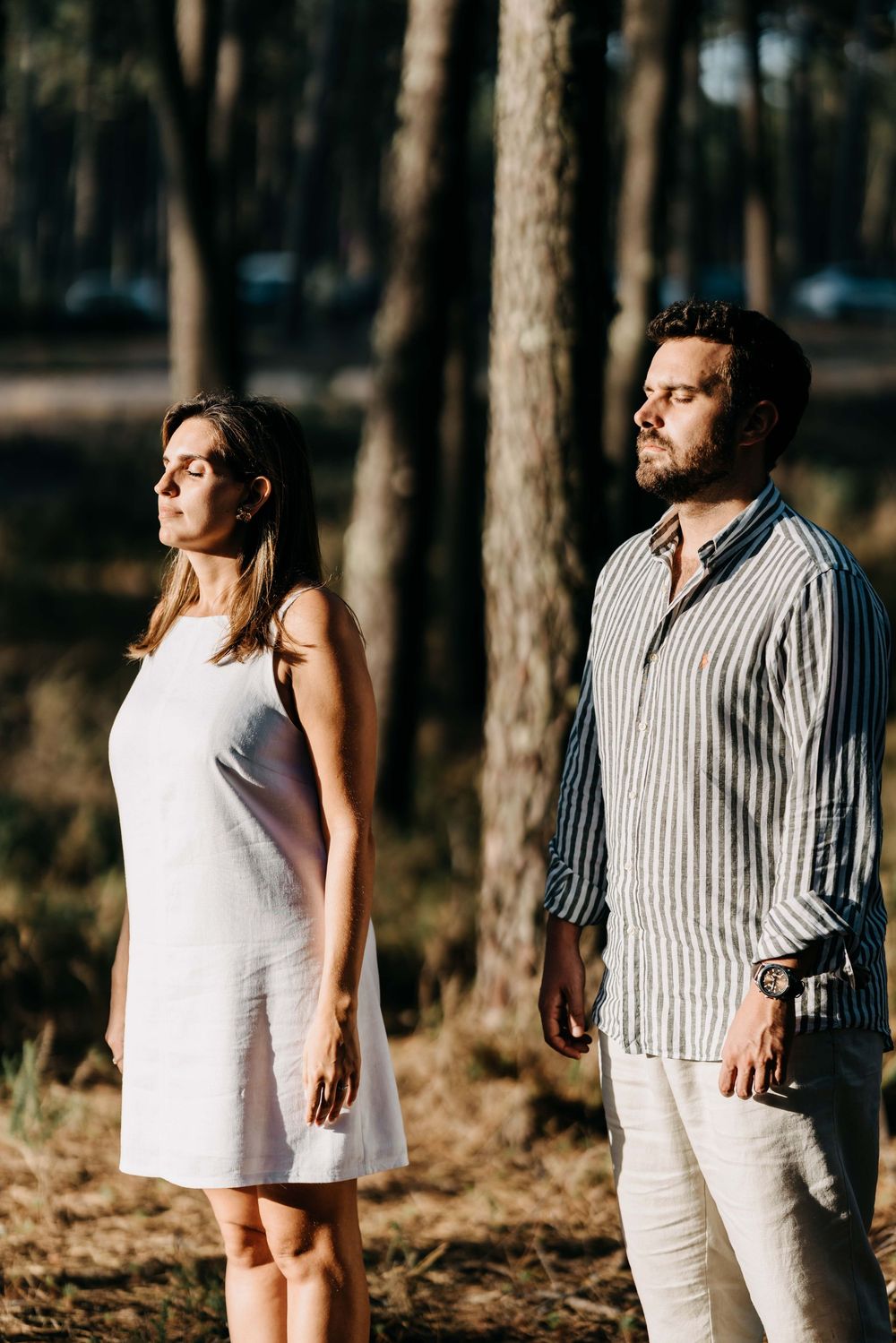 A couple in white and striped outfits poses together for romantic photos in a sunlit forest setting.