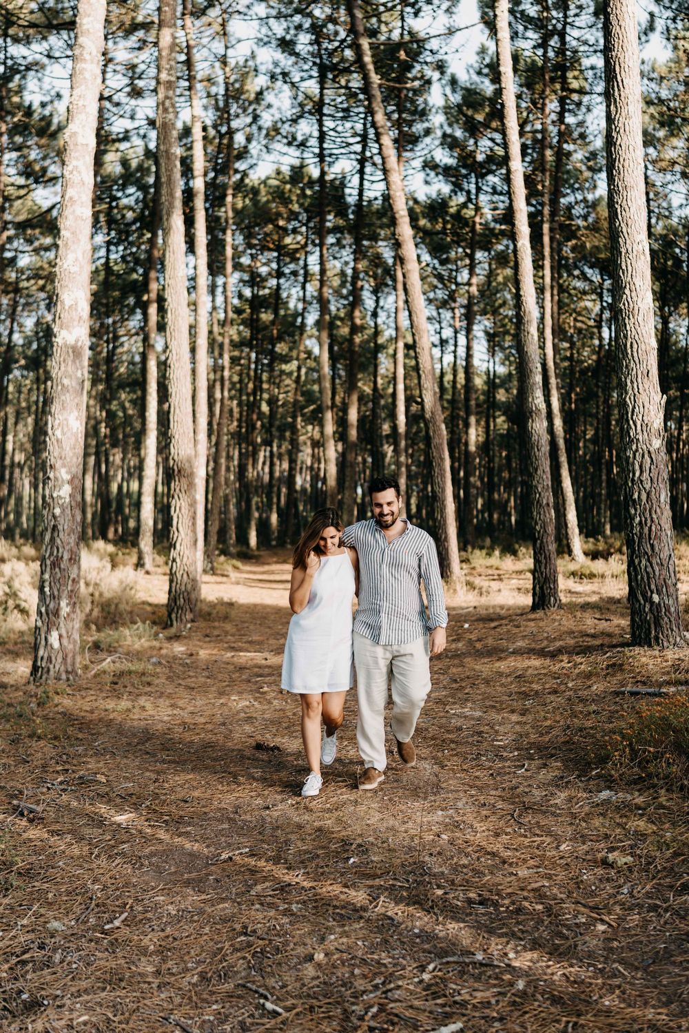 A couple walks hand in hand through a sunlit pine forest trail wearing coordinated white and beige outfits.