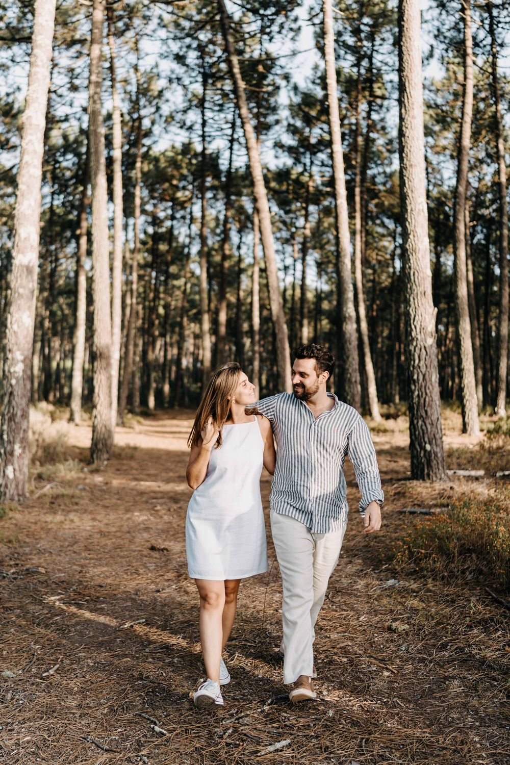 A couple in light summer outfits walking together through a sunlit pine forest on a warm afternoon.