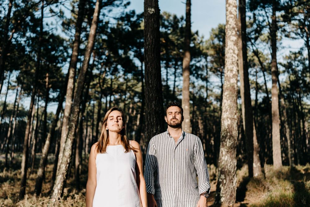A couple stands together in a sunlit pine forest wearing coordinated neutral-toned outfits.