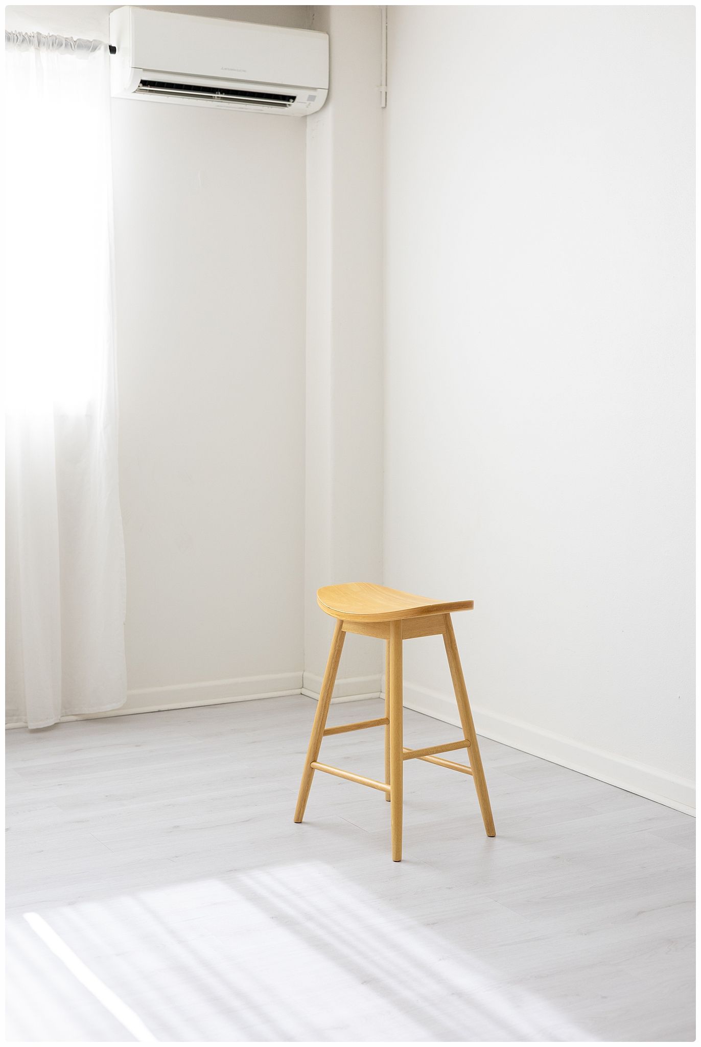 A minimalist wooden bar stool with natural finish stands on white floor beside a wall-mounted air conditioner.